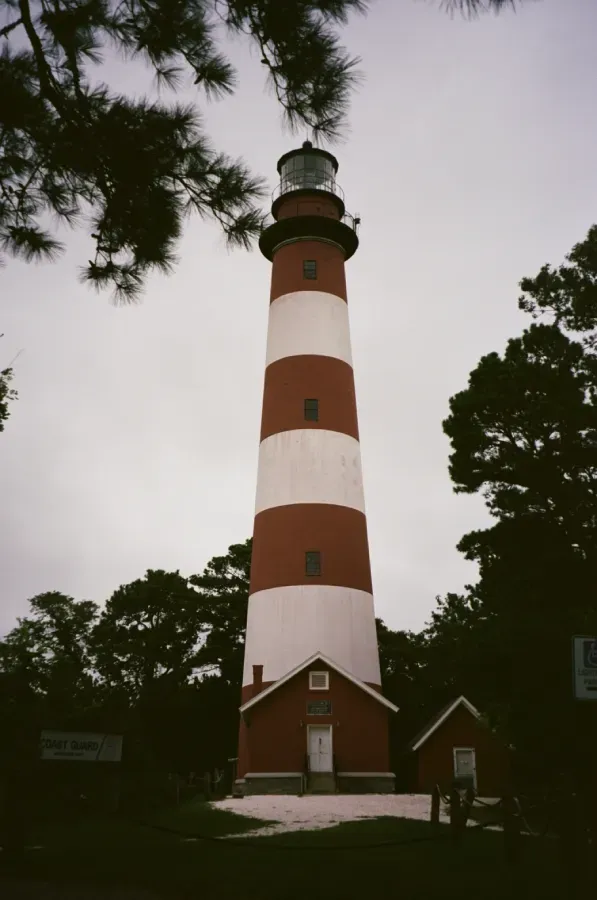 Assateague Island lighthouse with attached red buildings; surrounded by trees, overcast sky.