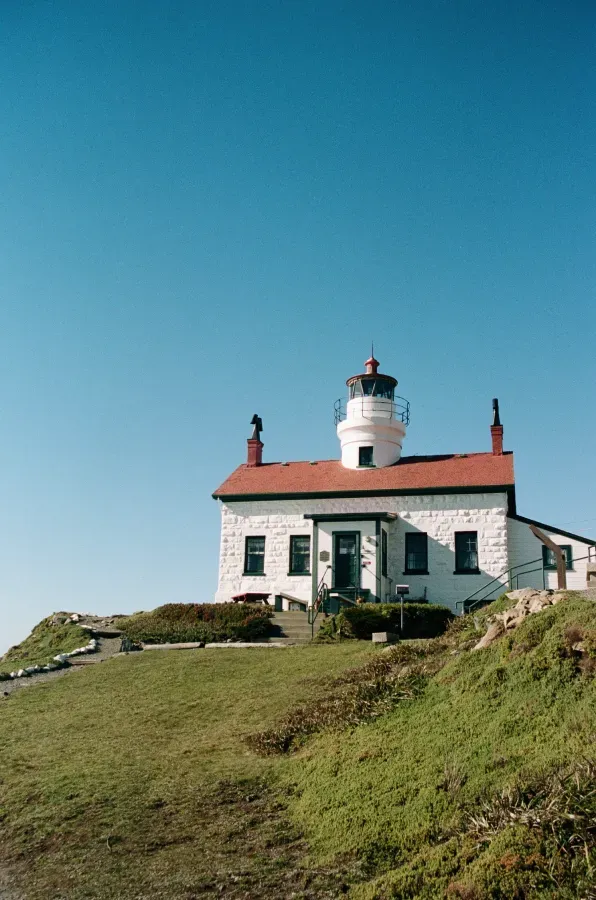 Charlee Remitz wearing sunglasses and a sweater smiles while sitting outdoors with a lighthouse and house in the background.