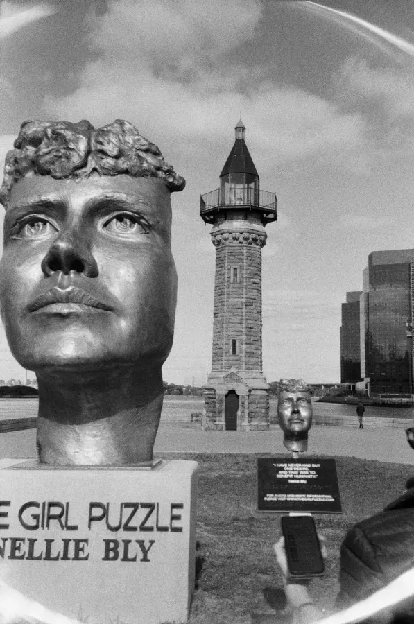 Black and white film photo: bronze bust of a person. Sign reads 