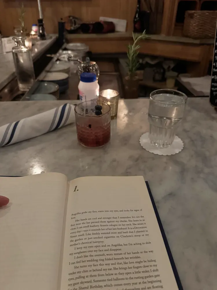 Open book on a bar counter beside A blueberry cocktail and a glass of water