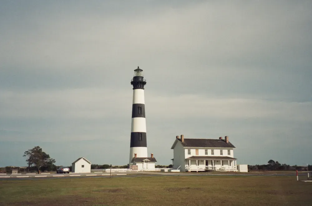 Bodie Island Lighthouse with its black and white stripes stands next to a historic two-story house on a grassy plain.