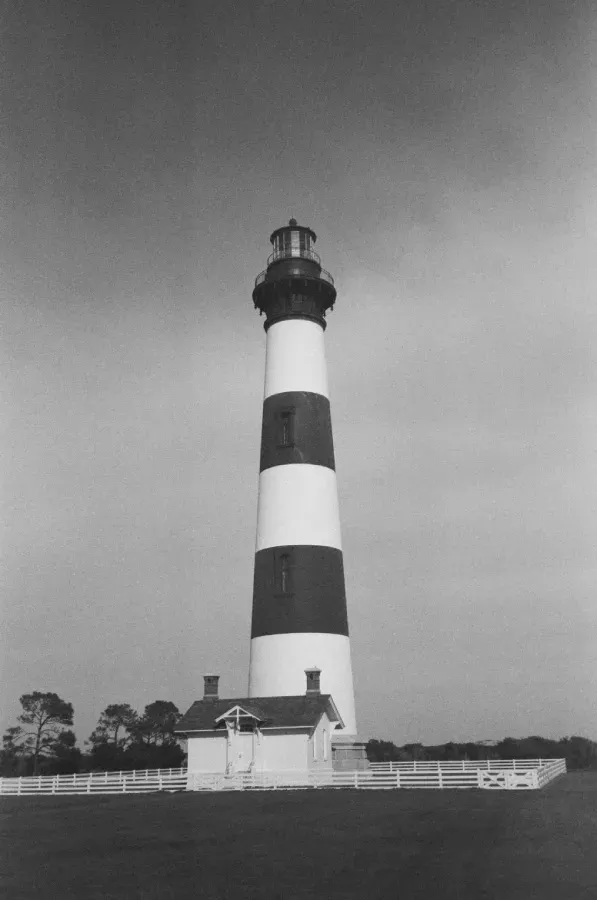 Black and white film photo of Bodie Island lighthouse, small white building at base, under a cloudy sky.