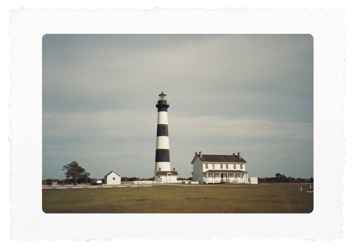 Black-and-white lighthouse beside a small white house on a flat grassy shoreline under a cloudy sky