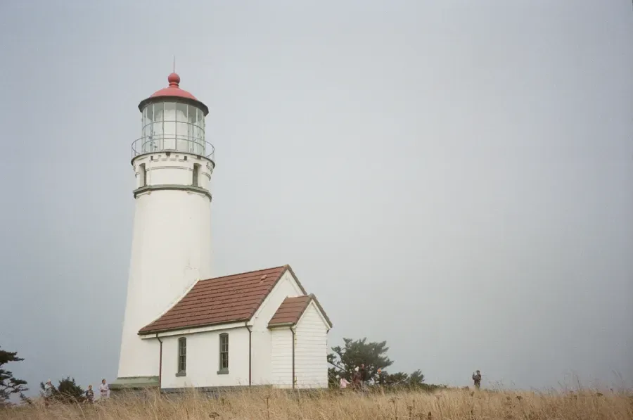 White lighthouse with red top and attached building on a grassy hill; overcast sky.