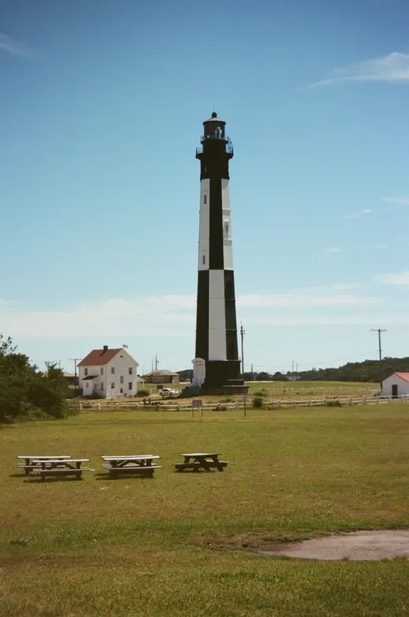 Cape Charles Black and white striped lighthouse on green lawn with picnic tables and small buildings under a blue sky.