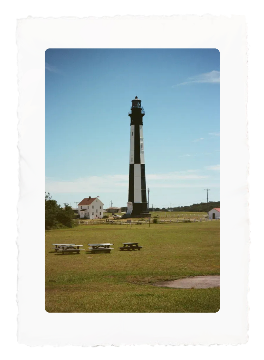 Tall black-and-white lighthouse on a grassy field under a blue sky, with a small house nearby.
