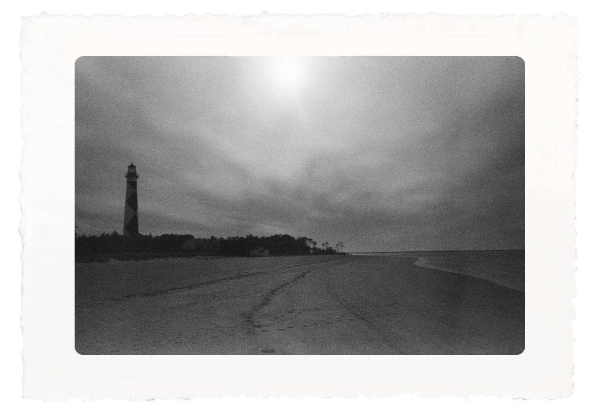 Black-and-white beach scene with a lighthouse and bright sun over the water