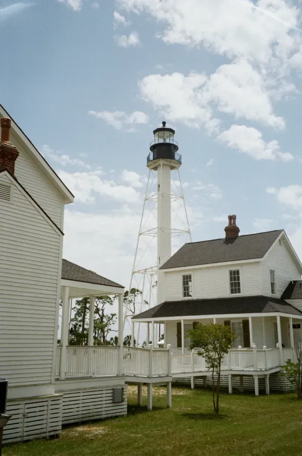 Cape San Blas lighthouse stands tall behind two white houses on a grassy lot, under a cloudy blue sky.