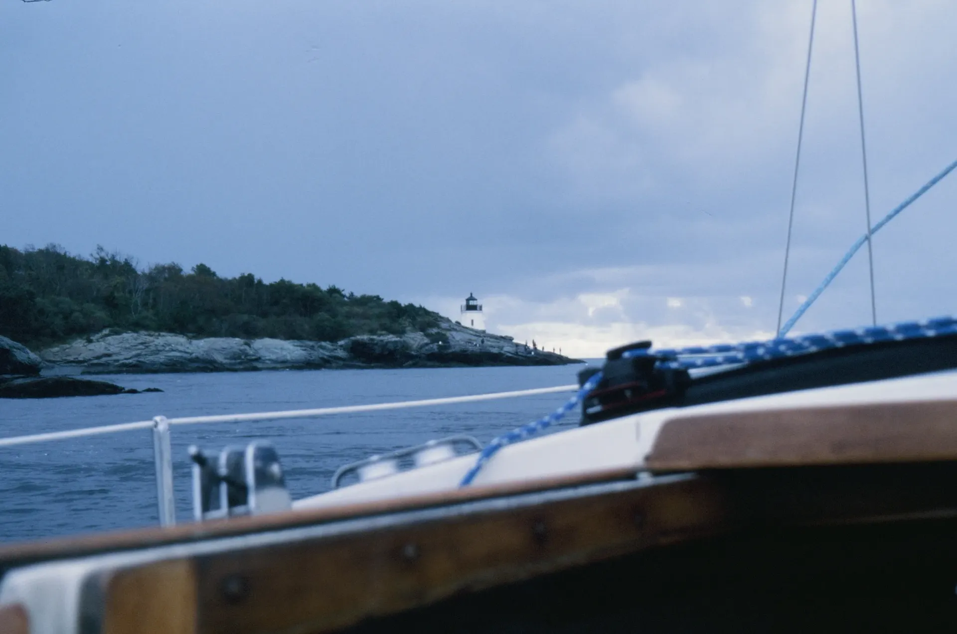 Sailboat view of a lighthouse on a rocky coast under a cloudy sky.