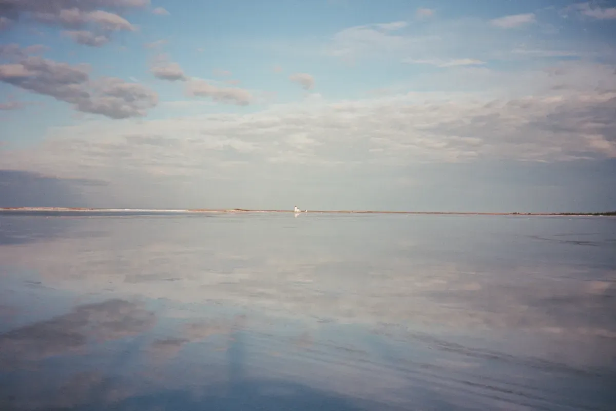 A mirror-like lake reflects a soft blue sky with wispy clouds and the Casumpec lighthouse in the distance