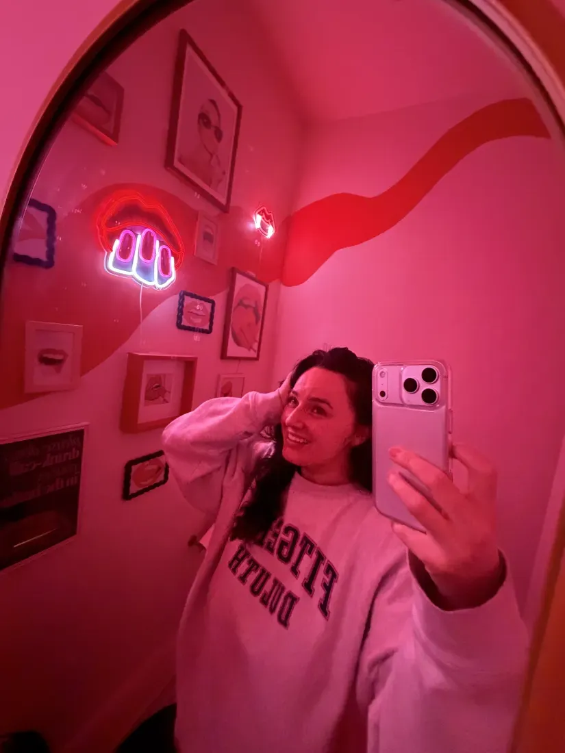 Charlee Remitz taking a mirror selfie in a red-lit room with framed wall art and a neon sign.