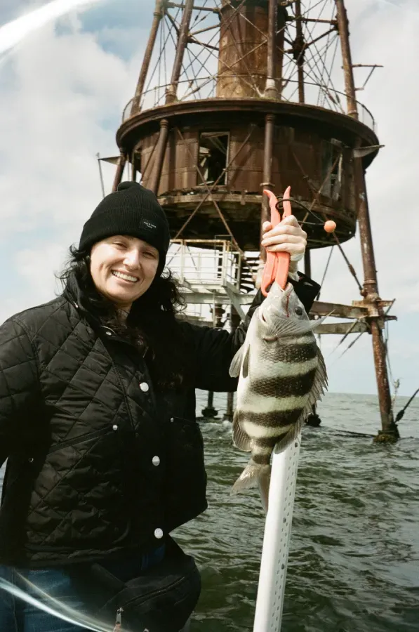 Charlee Remitz in a black jacket and beanie holds a striped fish with orange pliers in front of Ship Shoal lighthouse