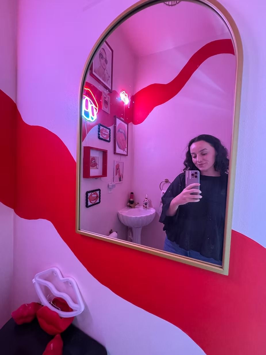 Charlee Remitz taking a mirror selfie in a pink-lit room with red wavy accents and artwork.