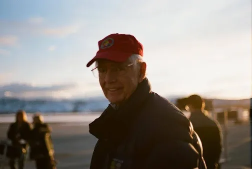 Grandpa Remitz standing on the shoreline smiling wearing a red cap and black jacket