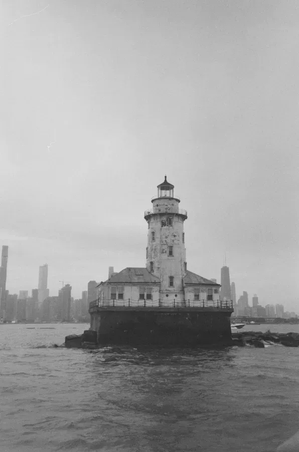 A weathered lighthouse on a rocky base in a body of water, city skyline in the background.