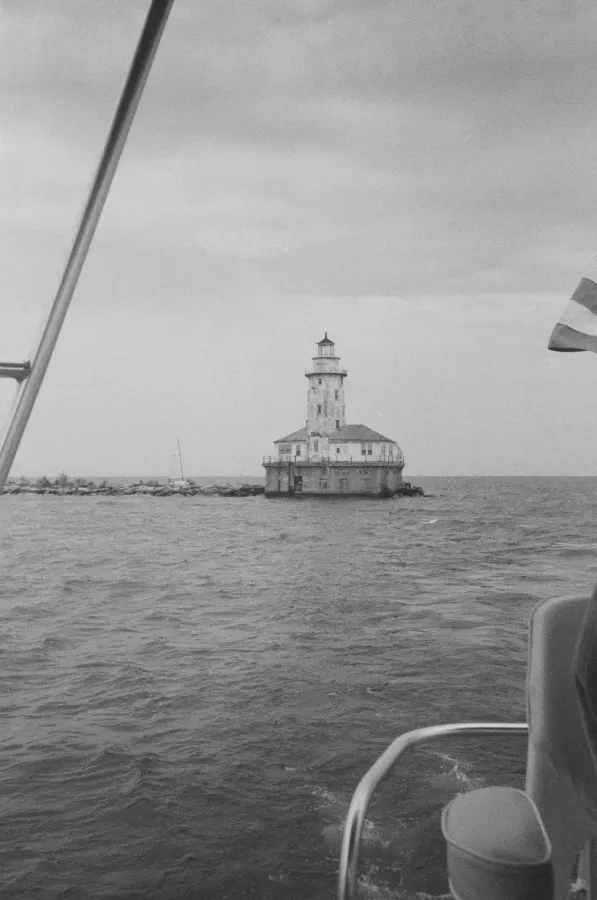 Chicago Harbor Lighthouse on a rocky breakwater in a body of water, viewed from a boat. Overcast sky.