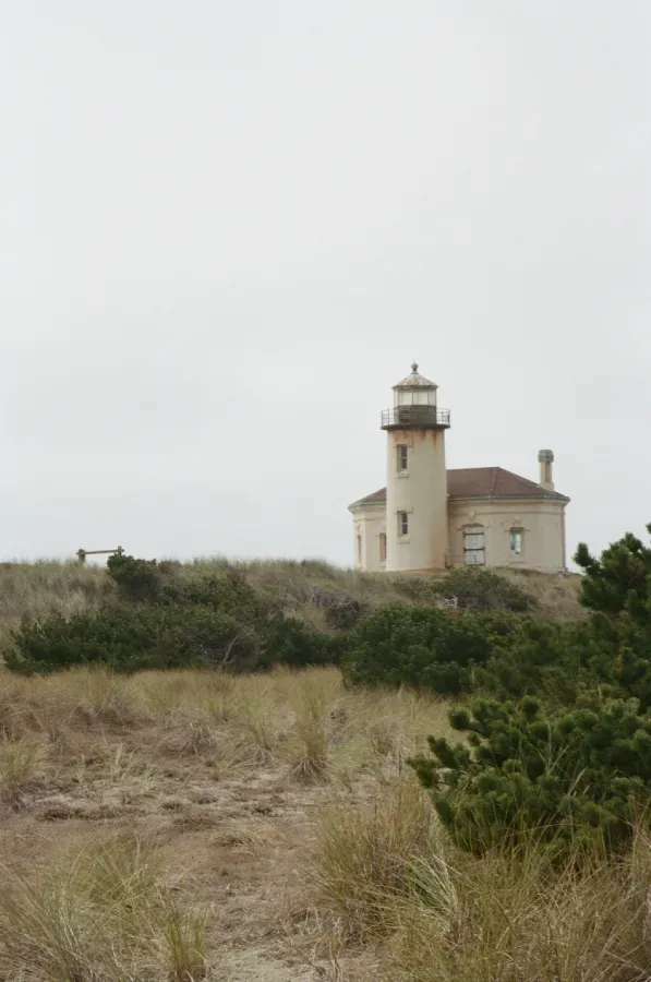 Coquille River Lighthouse on a dune, under overcast sky. There is a grey sky and shrubs in the foregrou