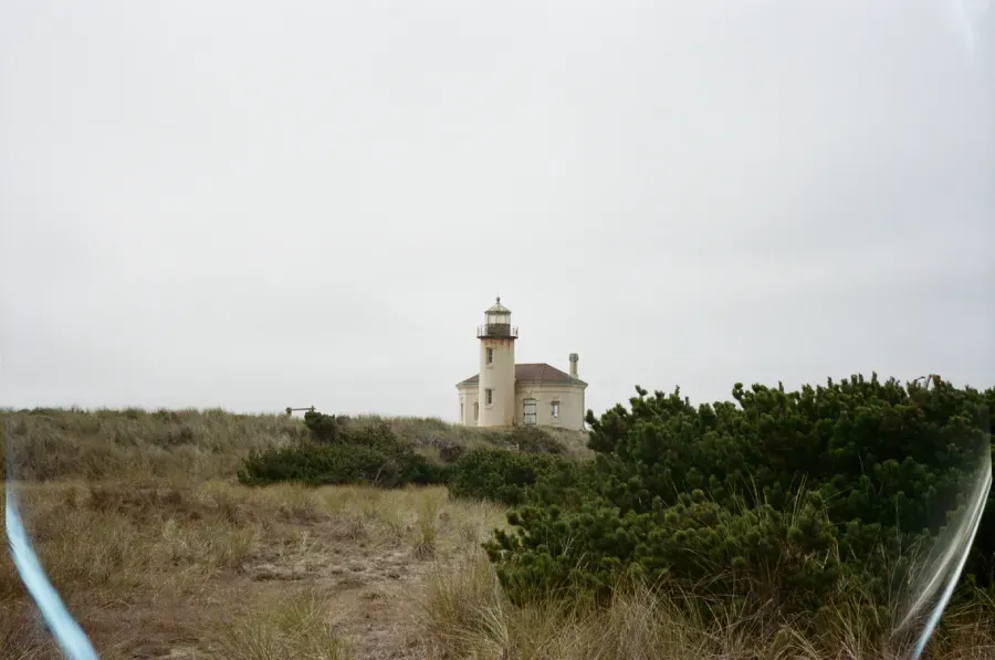 Film photograph of a Coquille River Lighthouse on a hill surrounded by coastal foliage under a cloudy sky.