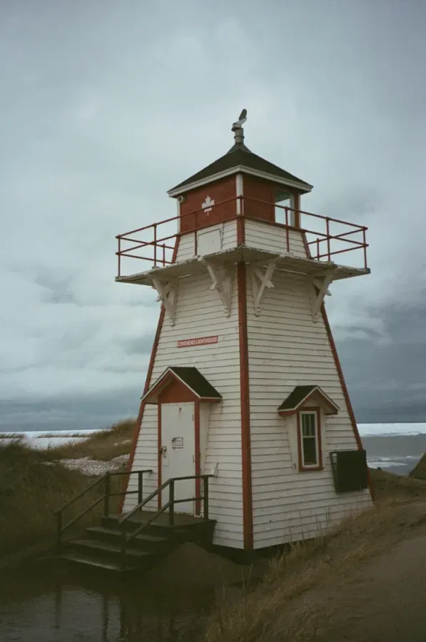 Covehead Harbour lighthouse with red trim and a railed lantern room stands on a dune under a cloudy sky.