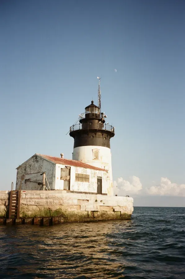 Detroit River Lighthouse on stone foundation with white and black tower, small attached building. Blue sky and water.