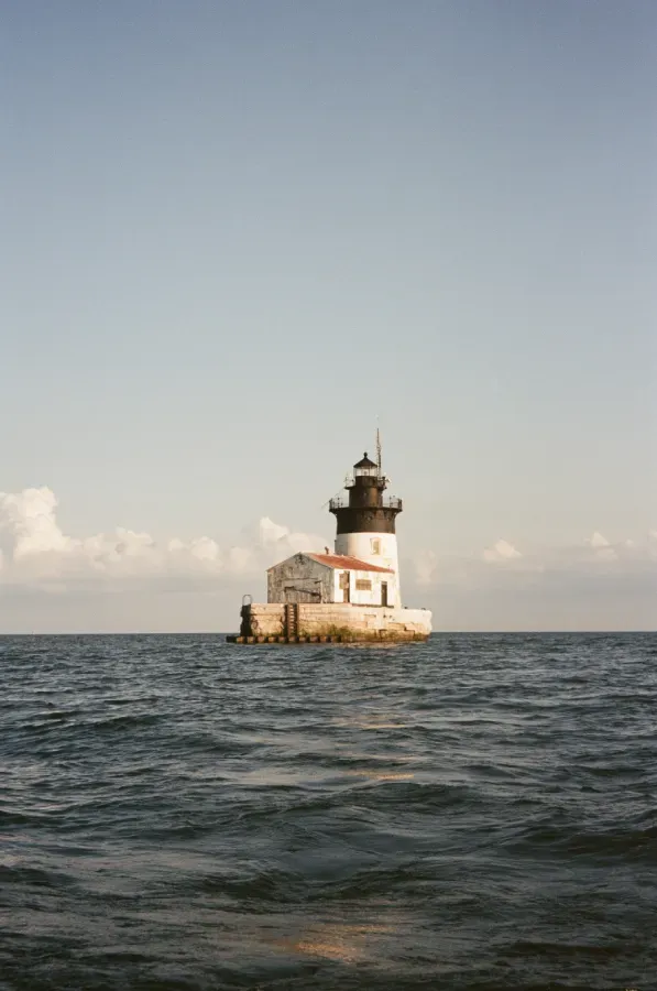 A person sits with legs crossed in front of a lighthouse and bushes under a clear blue sky.