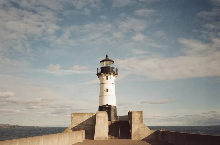A white lighthouse with a black top sits on a concrete pier against a partly cloudy sky and open water.