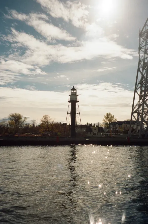 Duluth Harbor Lighthouse on a shoreline with a bridge in the background, under a bright sun and cloudy sky.