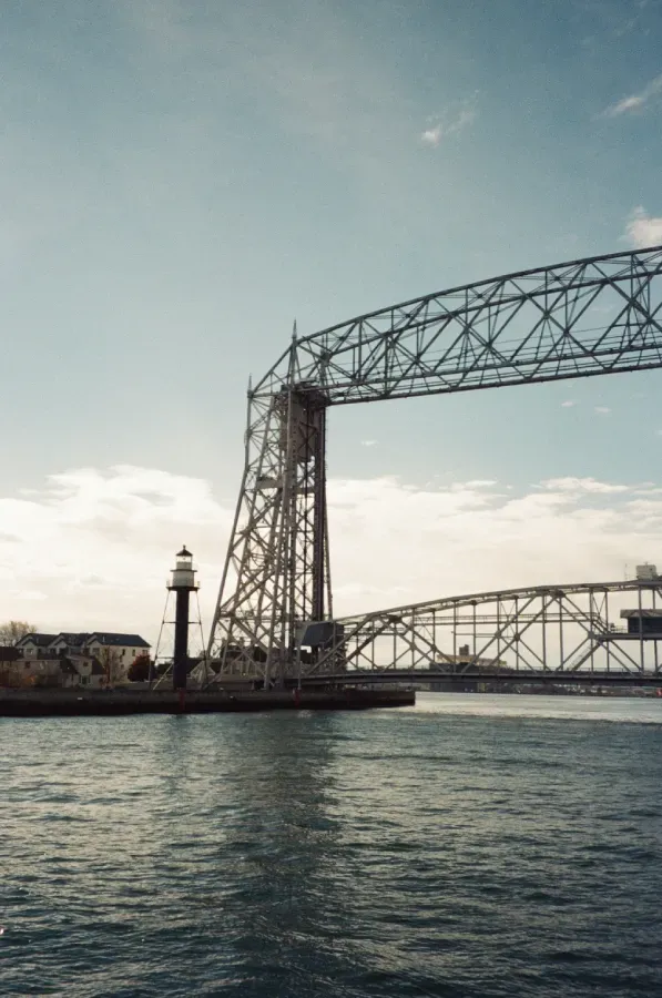 A tall, vertical lift bridge rises over blue water, with Duluth Harbor lighthouse and a second bridge visible.