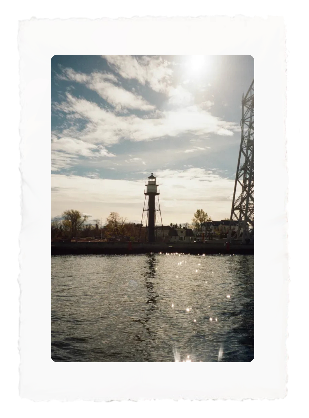 Lighthouse by a sparkling river under a cloudy sky, with a metal tower on the right and sunlit reflections.