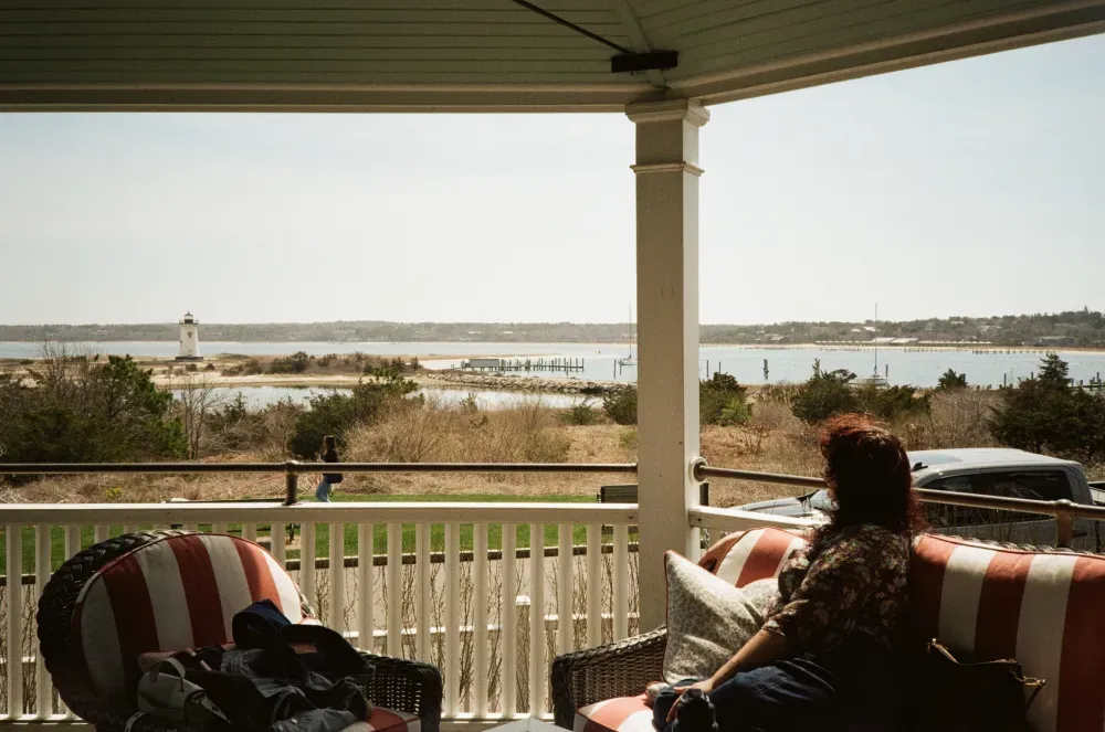 Covered porch overlooking a lighthouse on a waterfront, with a seated person and striped chairs in the foreground