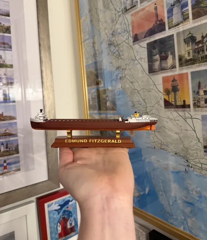 A hand holds a small wooden model ship labeled "Edmund Fitzgerald" in front of a framed wall map featuring lighthouses.