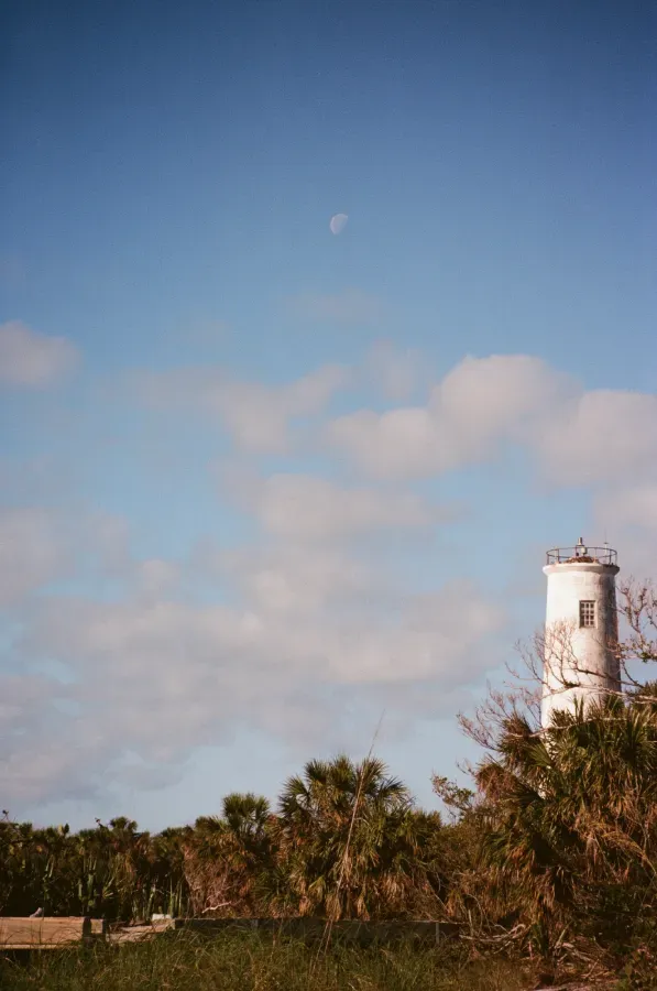 Egmont Key lighthouse on a grassy shore, under a blue sky with scattered clouds and a visible crescent moon.