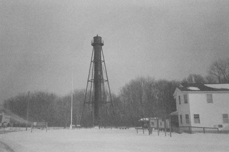 A black-and-white photo of Finns Point Range lighthouse standing in a snowy field next to a two-story building.