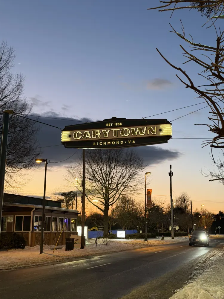 The Carytown sign glowing over a snowy street at dusk, with cars and bare trees against a pastel sky