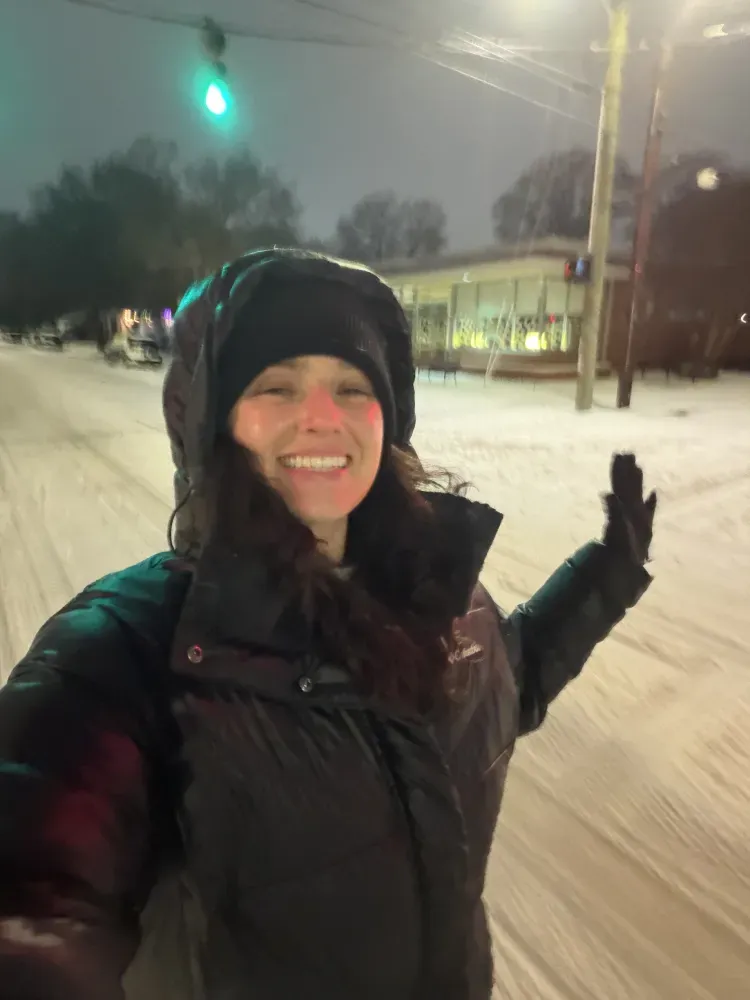 Charlee Remitz smiling in black winter coat and hat posing on a snowy street at night, waving under a green traffic light