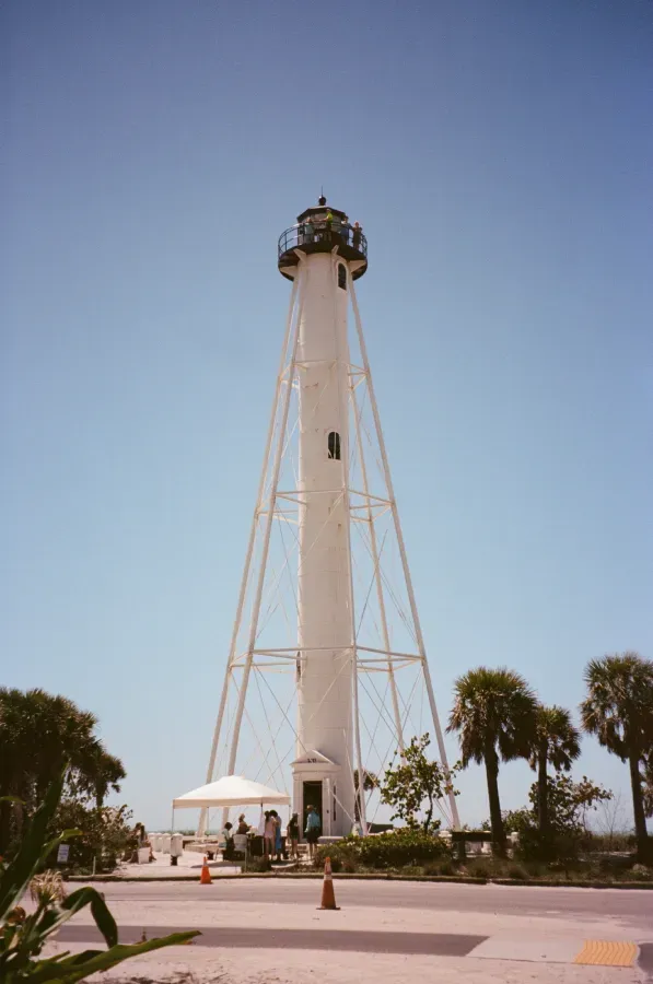 Gasparilla Island lighthouse against a blue sky, supported by metal frame. Palm trees and people near the base.