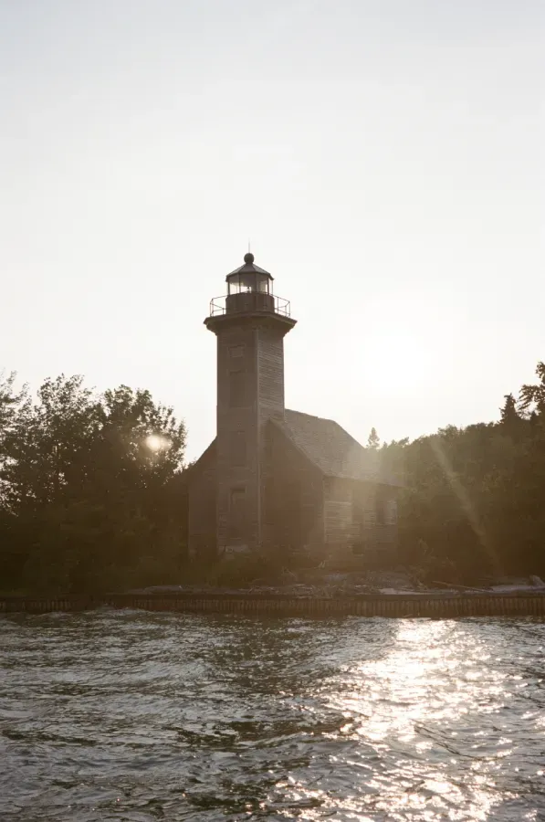 Grand Island Lighthouse at sunset on water surrounded by trees, sun rays reflecting on the water.