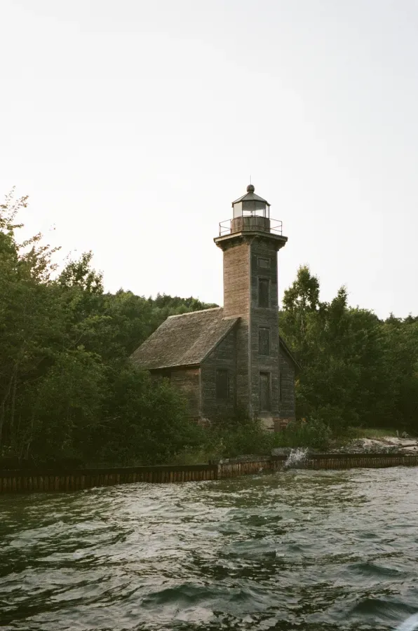 Grand Island East Channel lighthouse on a shoreline, surrounded by green trees. Water in the foreground.