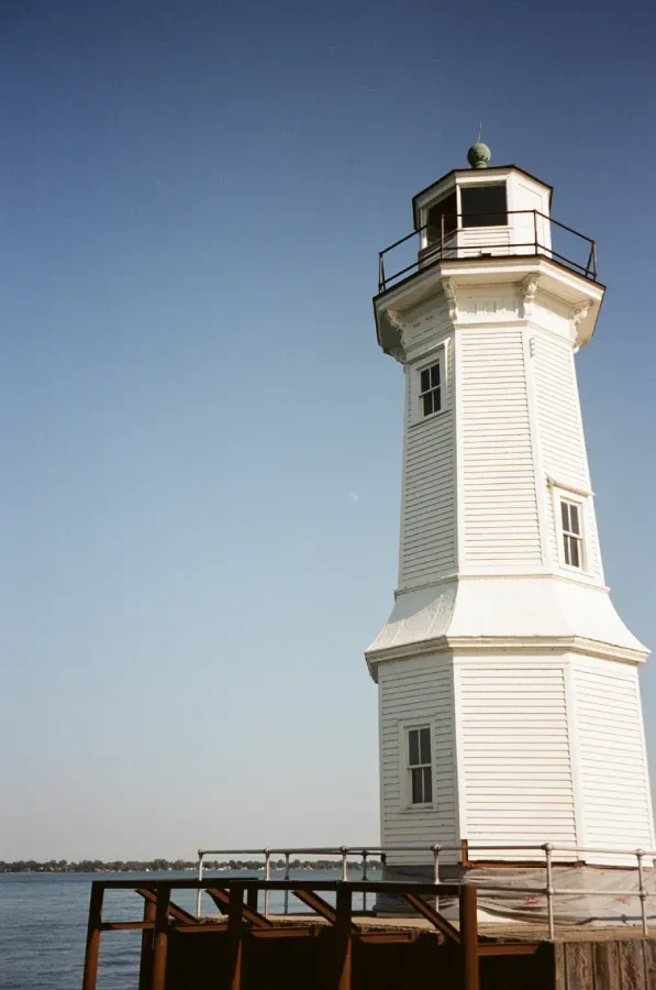 Grosse Ile lighthouse with black lantern room against a blue sky, standing on a wooden pier near water.