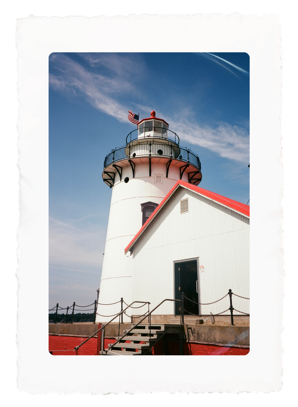 White lighthouse with red roof against a blue sky, viewed from below behind a fence