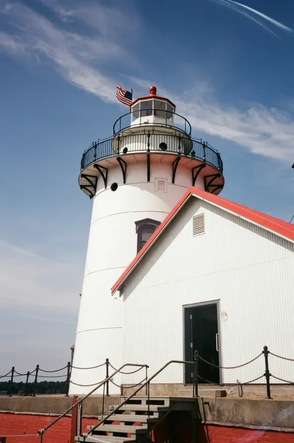 Harbor Beach lighthouse with red roof and trim under a blue sky, American flag on top.