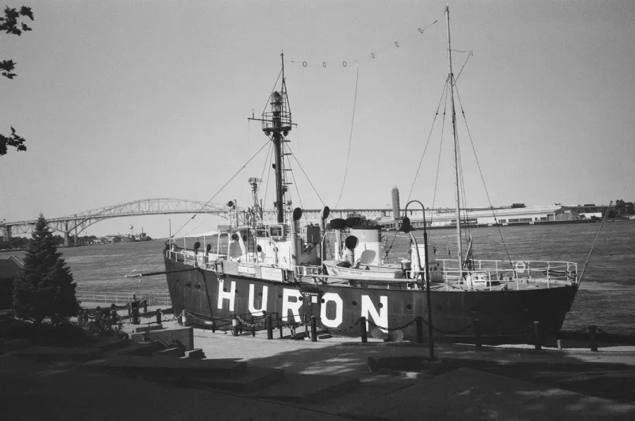 Black and white photo of Huron lightship docked on a waterfront. A bridge is in the background.