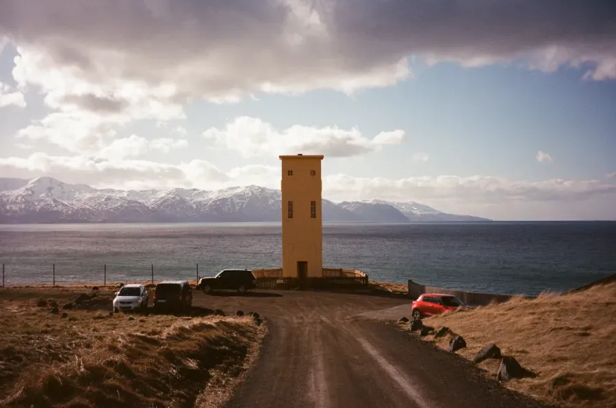 Husavik Lighthouse overlooking the coast with vehicles parked nearby. Mountains ocean and cloudy sky in the background.
