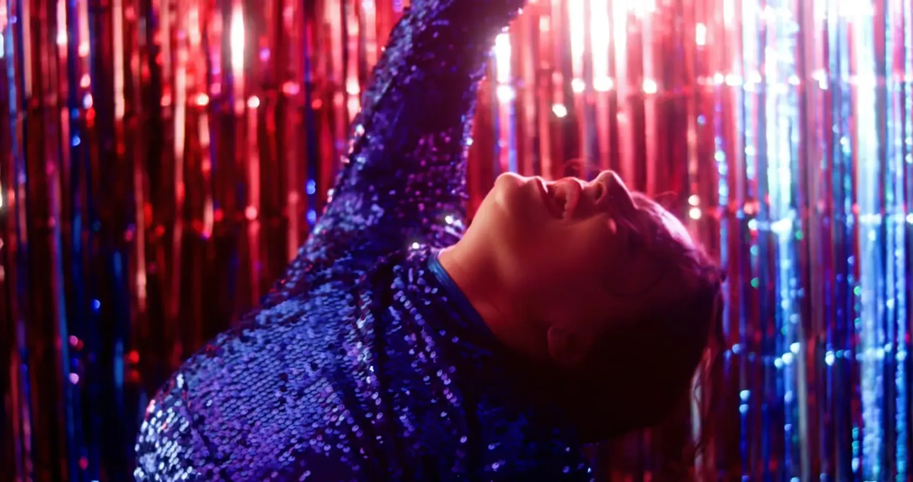 Charlee Remitz n a blue sequined outfit raises arm, looking up, in front of a shiny red and blue backdrop.
