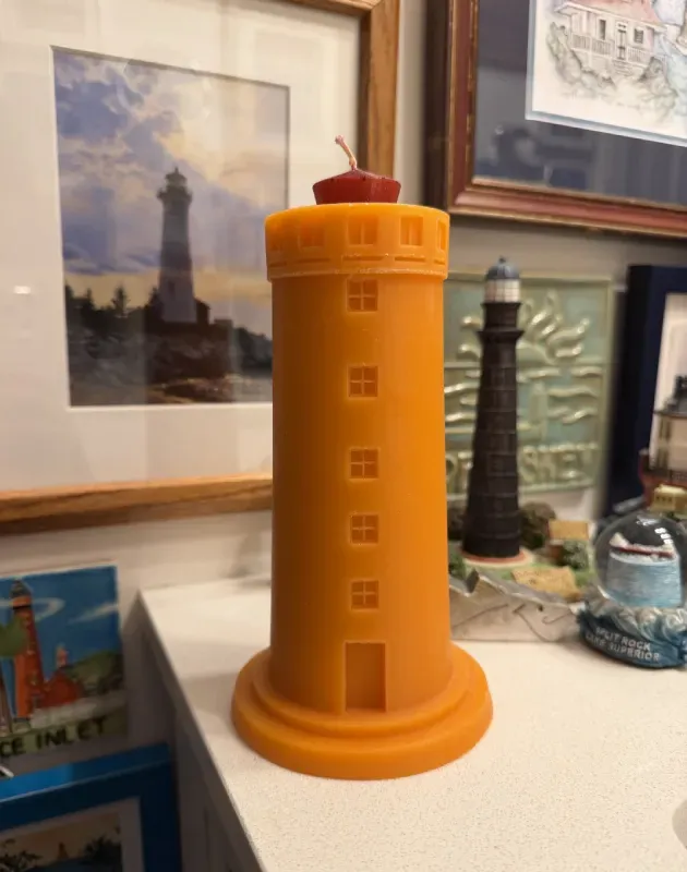 A yellow, lighthouse-shaped candle sits on a surface surrounded by other lighthouse-themed decor and framed pictures.