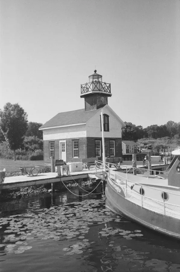 Black and white film photo of Kalamazoo lighthouse building with a boat docked on a lake.