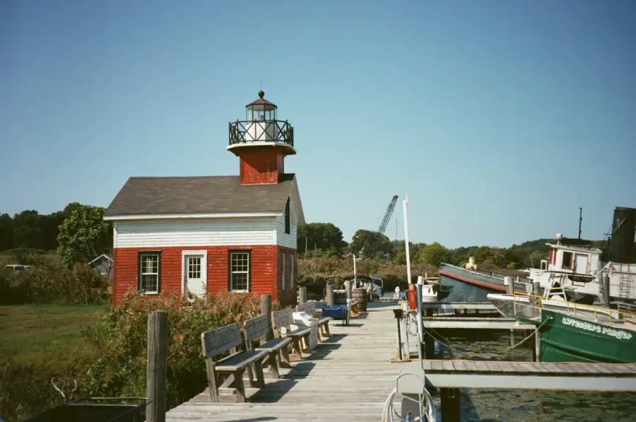 Red and white Kalamazoo lighthouse with black roof sits on a pier with benches and boats on a sunny day.