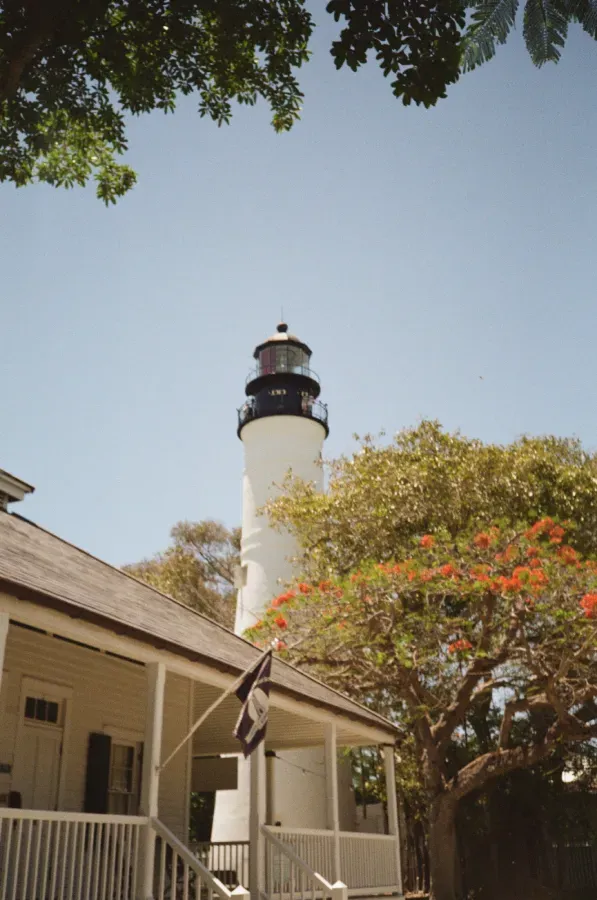 Key West lighthouse next to a yellow house with a porch, under a clear blue sky, framed by green and orange foliage.
