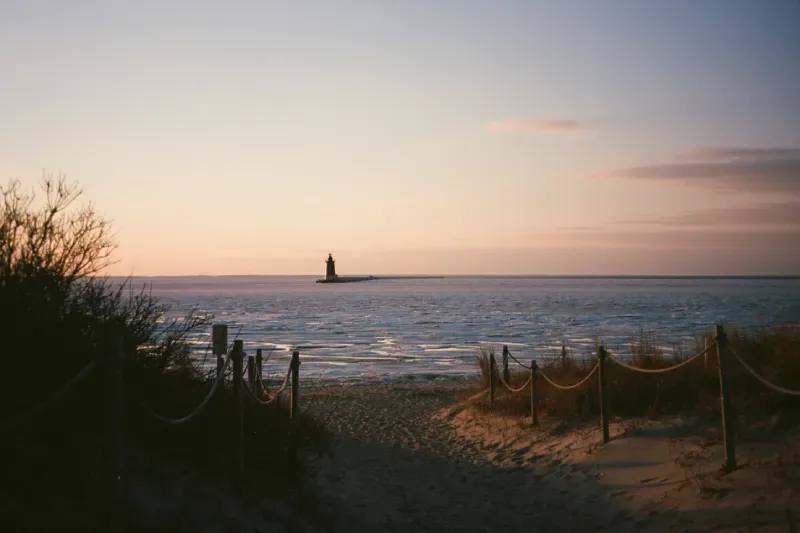 A sunset view over a sandy path leading to the ocean, with Lewes Breakwater lighthouse in the distant horizon