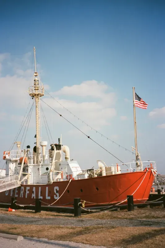 The red lightship Overfalls is moored at a dock, featuring a tall mast, white superstructure, and a flying U.S. flag.